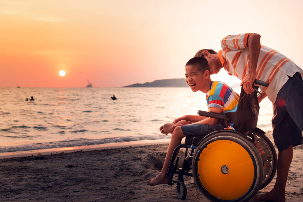 Boy in wheelchair being pushed by older boy on the beach sunsetting